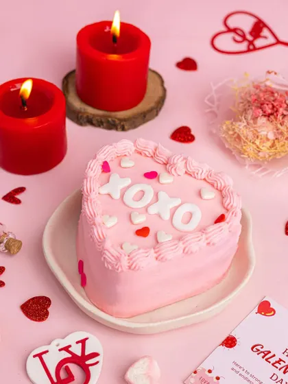 A cute pink heart-shaped bento cake with "XOXO" written on it, styled with red candles for a romantic Valentine's vibe.