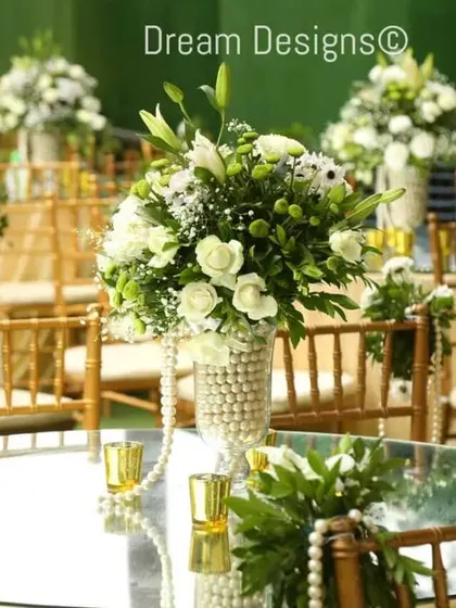 An elegant table setup featuring a mirrored tabletop that reflects the beautiful centerpiece. The vase is adorned with pearls, and the chairs are decorated with pearl strings for a touch of classic glamour.