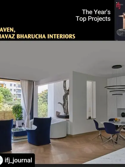 This image shows the dining and lounge area of 'The Haven,' which opens onto a balcony with green views. The design is minimal and clean, using a neutral palette with pops of deep blue in the chairs to create a modern and airy space.