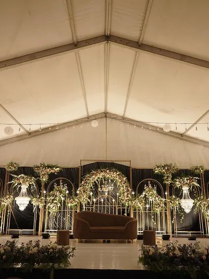 We designed this elegant reception stage inside a tented structure, proving that any space can be transformed. The dark backdrop makes the gold framework, chandeliers, and white floral arrangements pop, creating a sophisticated and intimate ambiance.