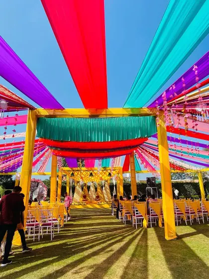 A vibrant outdoor Haldi or Mehendi function with a massive tent made of colorful drapes in shades of red, purple, blue, and yellow. This creates a lively and celebratory atmosphere for a large crowd.