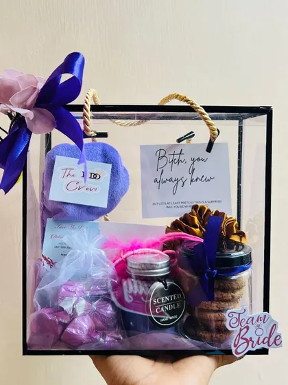 A hand holding a single bridesmaid hamper, showcasing its design. The purple ribbon and flower add an elegant finishing touch.