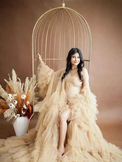A glamorous and confident portrait. The mom-to-be is seated in a golden birdcage chair, wearing a stunning ruffled nude gown.