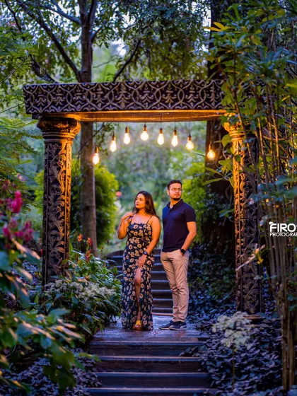 A stylish and moody night shot on a wooden pathway. The hanging Edison bulbs and surrounding greenery create a cinematic and slightly mysterious atmosphere.