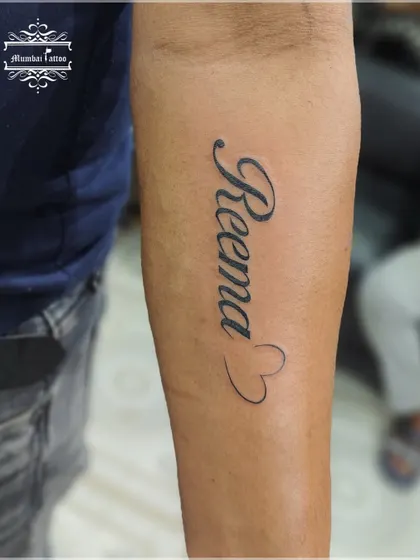 The name "Reema" ending in a heart. This is a classic and popular style for name tattoos, and we execute it with clean, elegant lines.