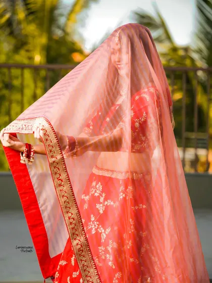 The bride playfully holds her sheer dupatta, creating a beautiful silhouette against the bright outdoor light.