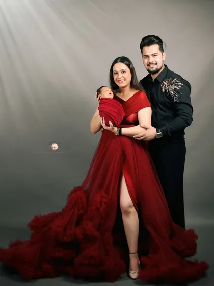A glamorous family portrait celebrating a new beginning. The parents smile warmly while holding their newborn, with the mother's stunning red gown creating a bold and beautiful statement in the studio.