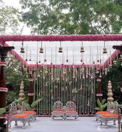 A warm and inviting mandap design featuring pillars adorned with pink flowers and a canopy of hanging floral strings and traditional bells. This setup at The Tamarind Tree's amphitheater creates an intimate and sacred space for the wedding rituals.