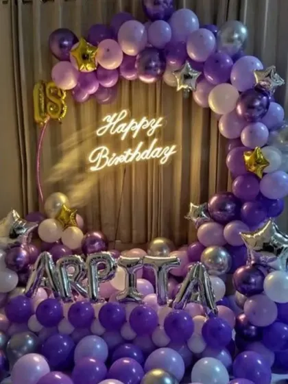 A rich purple and silver theme for an 18th birthday. This home setup includes a balloon arch, foil balloons for the name "ARPITA", and a neon "Happy Birthday" sign, creating a festive focal point in the room.