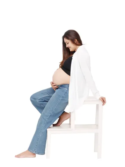 A relaxed, full-length portrait of a mom-to-be in jeans and a white cardigan, showing off her bare bump. The clean white background and simple prop create a modern and minimalist feel.
