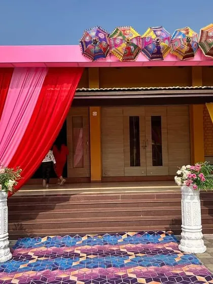 A colorful entrance gate decoration for a wedding event. The pink frame is draped with red and pink fabric and topped with decorative Rajasthani umbrellas for a festive welcome.