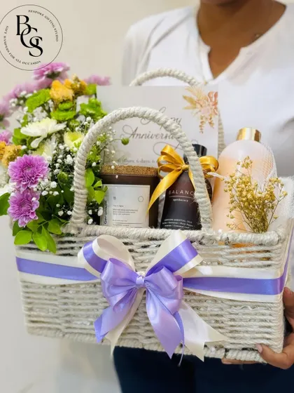 An elegant anniversary hamper in a white woven basket. It's filled with fresh purple and white flowers, scented candles, and bath products, creating a relaxing and romantic gift for a special couple.