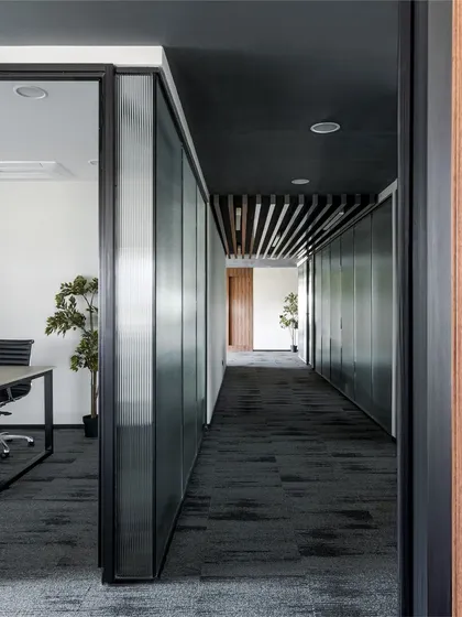 A view down an office corridor, showcasing the interplay of materials. Fluted glass panels on one side contrast with dark, solid doors on the other, while a dark ceiling and patterned carpet guide the way.