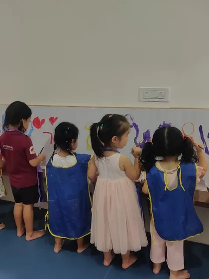 A line of children in their aprons, fully immersed in painting a large canvas on the wall. This is what joyful, messy learning looks like.