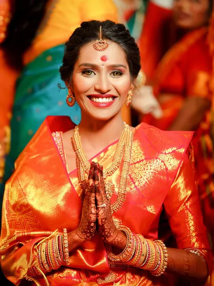 A vibrant portrait of the bride in a stunning orange silk saree. With her hands joined in a welcoming 'namaste', her joyful smile lights up the frame.