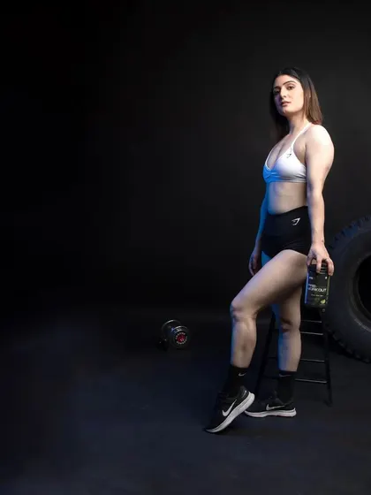 A side profile of a female model in a strong pose, holding a supplement bottle. The dramatic lighting highlights her physique and the product.