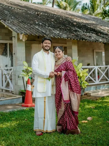 A lovely portrait of a South Indian couple in their traditional wedding attire, standing outside a charming cottage.