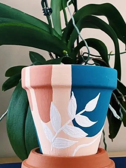 A close-up of a hand-painted planter with a bold, leafy design. The contrast between the terracotta, blue, and white makes the plant inside truly stand out.