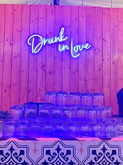 Two of our bartenders ready for a wedding party behind a custom bar. The "Drunk in Love" neon sign adds a fun, romantic touch to the setup.