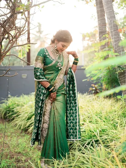 A full-length shot in a serene garden setting. The bride's green saree and soft makeup look harmonious and beautiful against the natural backdrop.