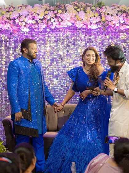 The couple on stage during their sangeet, interacting with the host. The backdrop of shimmering sequins and a floral halo creates a glamorous setting for the evening's festivities.