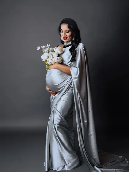 A full-length studio shot of an expectant mother in a graceful silver saree, holding a bouquet of white peonies. The long, flowing drape of the saree adds a touch of regal elegance to the portrait.