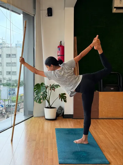 A student practices Natarajasana (Dancer's Pose) using a stick for balance. This prop helps maintain stability while focusing on the deep backbend and stretch.