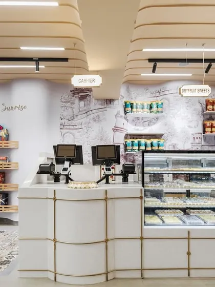The billing and display counter at Sangam Sweets features metal details inspired by traditional column designs. The curved counter showcases a wide range of Pan-Indian sweets against a backdrop of historical artwork.