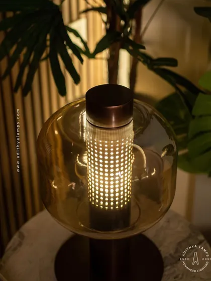 This table lamp features a perforated inner cylinder shielded by an outer amber glass shade. The design creates a mesmerizing pattern of light, offering a warm and intricate glow for a side table or console.