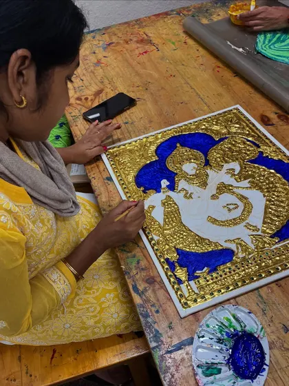 A student meticulously applies a deep blue background to her Tanjore painting of Radha and Krishna, making the gold relief work stand out beautifully.