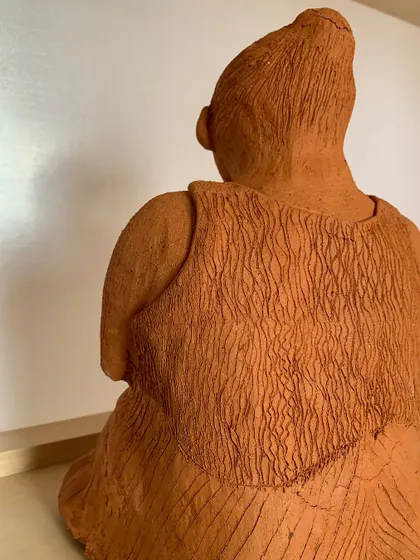 A back view of the seated woman sculpture, showing the intricate texture work on her clothing. Every mark is made by hand, adding to the piece's unique character.