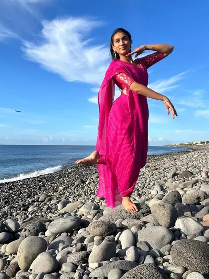 A dance pose in a bright pink saree on a pebble beach. The contrast between the vibrant color of the saree and the blue sky creates a visually striking image for fashion and travel campaigns.