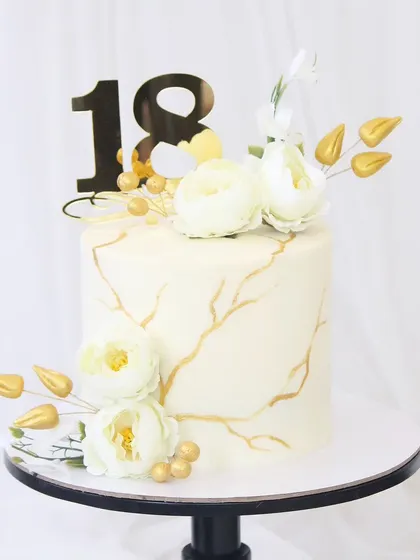 An elegant cake for an 18th birthday, featuring a white marble effect with golden veins. The design is completed with delicate white sugar flowers and gilded leaves.