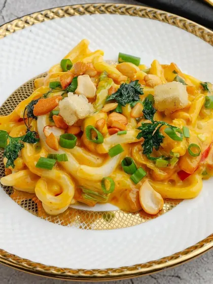 A close-up of our Peruvian Udon Noodles. The thick, chewy noodles are coated in a vibrant yellow sauce and topped with crunchy peanuts and fresh spring onions for a multi-textured dish.