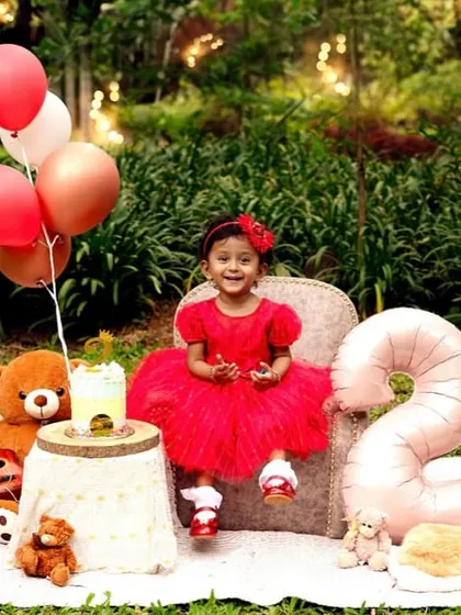 A happy smile from the birthday girl. The vibrant red dress stands out beautifully against the lush green background of the park.