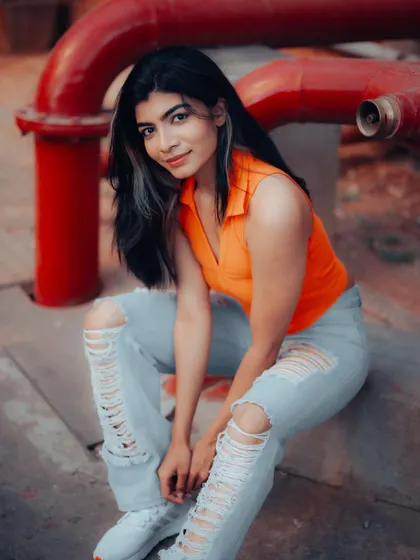 A relaxed portrait of a model sitting in an industrial area. The contrast between her bright orange top and the red pipes creates a vibrant and edgy look.