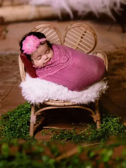 A full-length view of a newborn sleeping peacefully on a small cane chair, surrounded by greenery.