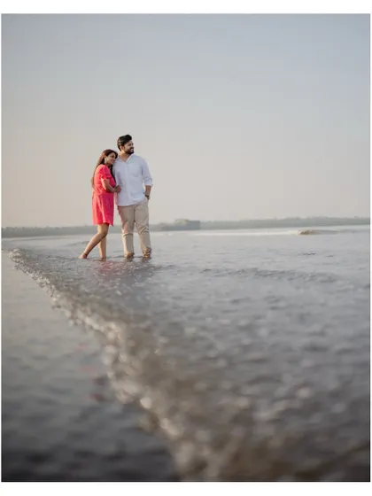 The couple stands at the water's edge on the beach, looking out at the horizon. A serene and thoughtful moment from their pre-wedding shoot.