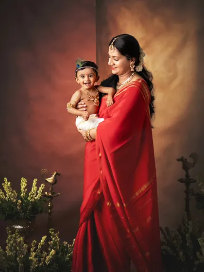 A mother in a striking red saree holds her baby, who is dressed as little Krishna. The rich colors and classic posing give this photo a beautiful, painterly quality.