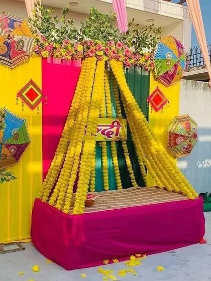 A canopy-style seating for a Haldi ceremony, created with strings of marigold flowers over a pink diwan. The backdrop is a mix of bright yellow, pink, and green drapes.