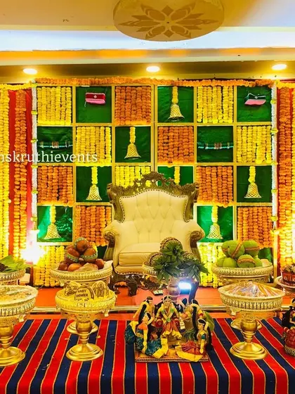 This traditional setup features a checkered backdrop in green and yellow, with hanging bells and marigold garlands. The striped floor mat adds to the classic, festive feel.