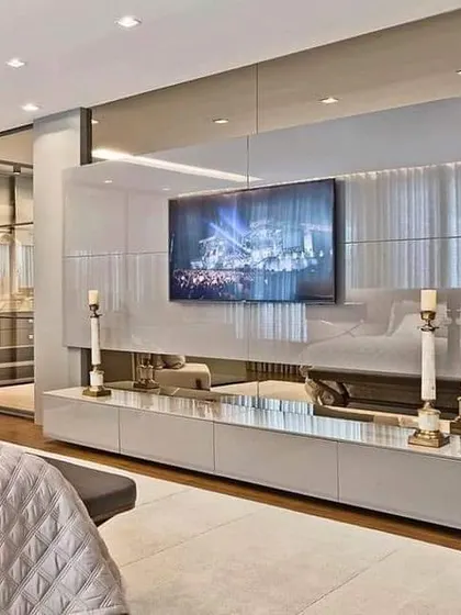 This bedroom TV unit is built into a mirrored wall, which makes the room feel incredibly spacious and bright. The glossy white console reflects the light, adding to the glamorous and airy feel.