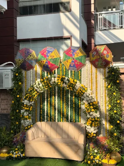 A beautiful outdoor Haldi setup featuring a floral arch of yellow and white marigolds against a backdrop of green and white striped drapes. The scene is brightened by colorful Rajasthani umbrellas suspended from above.