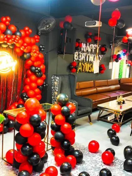 A dramatic red and black themed birthday setup in a private party room. The decor includes a balloon arch with a fringe curtain and balloon pillars.