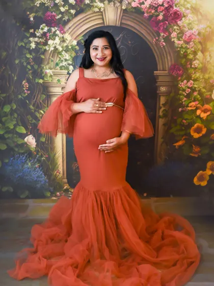 A beautiful studio portrait of a mom-to-be in a rust-colored gown, cradling her baby bump in front of a floral arch.