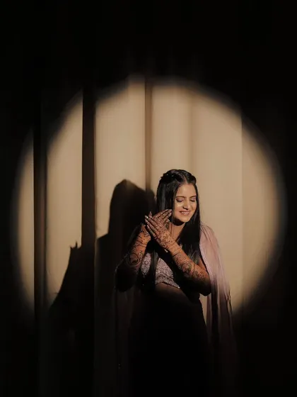 An artistic shot of a bride in a spotlight, her hands joined in a graceful pose. The shadows and light play beautifully on her mehandi.