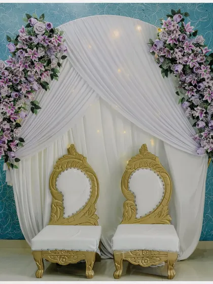 The elegant stage setup for an engagement ceremony. The white and gold chairs against a backdrop of white drapes and purple flowers create a regal look.