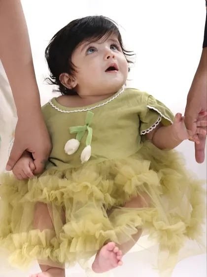 A baby girl in a lovely green dress looks up with wonder while holding her parents' hands.