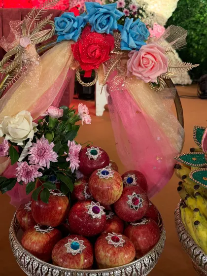 A silver bowl of fresh apples, each decorated with a small, sparkling bindi. The platter is adorned with a colorful floral arrangement, making it a feast for the eyes.