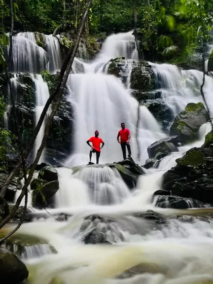 A long-exposure shot of two trekkers standing on rocks in a waterfall, creating a silky, dreamlike effect with the water.